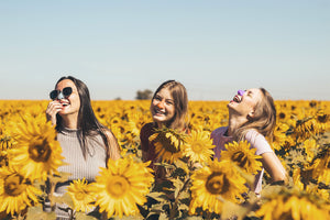 Three girls smiling in a sunflower field each with a different colored Nöz sunscreen on their nose.